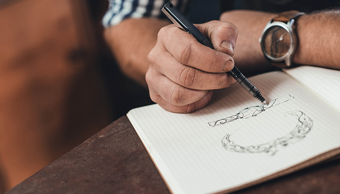 Closeup of a jeweler leaning on a bench sketching out new jewelry designs in a notebook while working in his shop